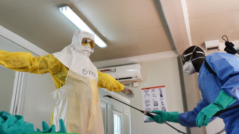 A health worker is doffing the personal protection suit he has been wearing. There are specific protocols for donning and doffing in the context of Ebola to ensure the health worker is not infected. For every layer of clothes, a sprayer is disinfecting the health worker with a chlorine solution.
